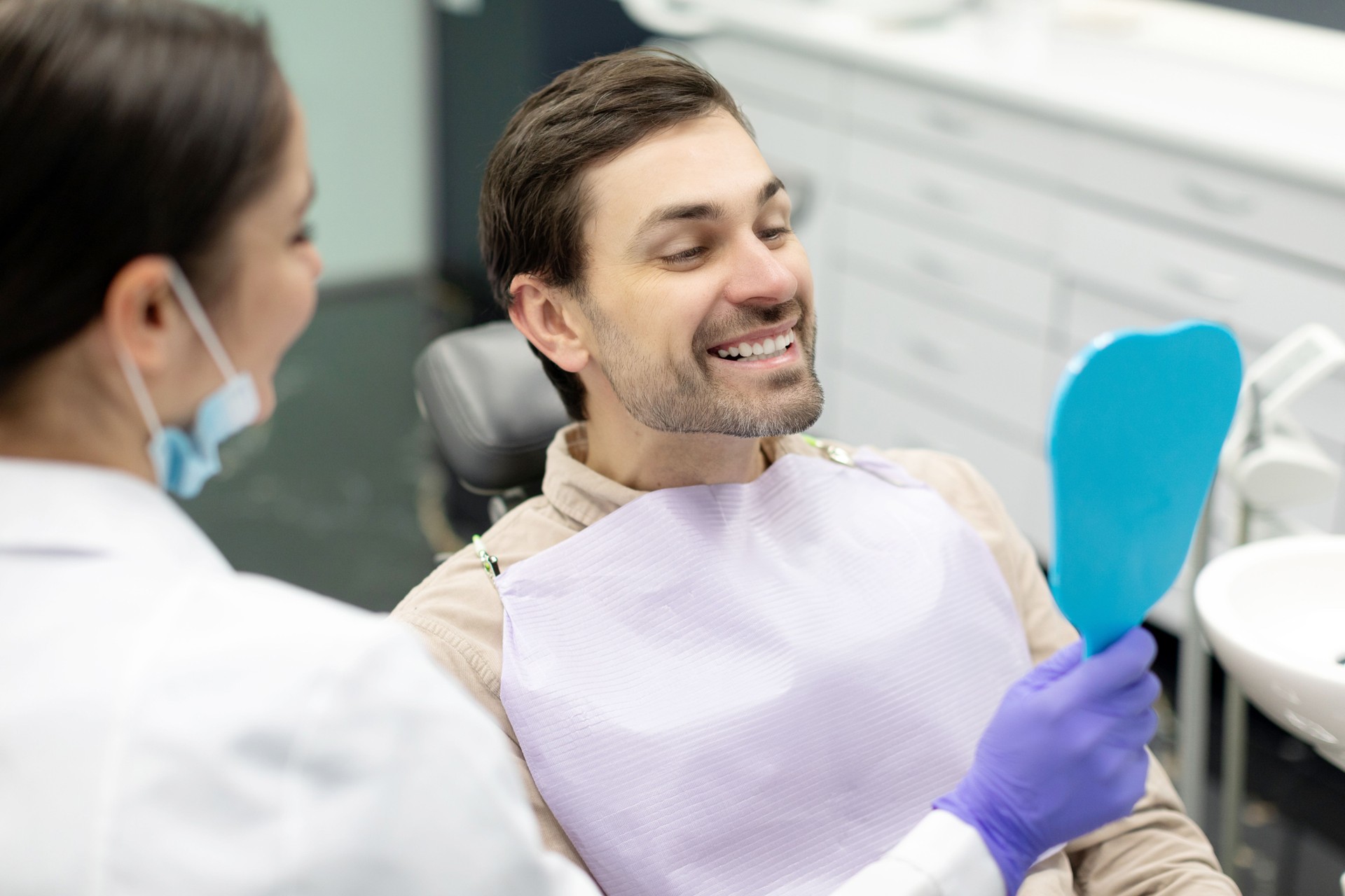 Happy man looking at mirror after teeth treatment in modern clinic, male patient sitting in chair in stomatological cabinet and enjoying his new smile